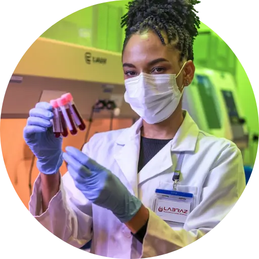 Uma profissional de laboratório segurando tubos de ensaio com amostras de sangue em um ambiente laboratorial. Ela está usando um jaleco branco, máscara facial, luvas azuis e um crachá de identificação com o logotipo "Labraz". Ao fundo, há equipamentos de laboratório, e a iluminação cria um tom esverdeado, indicando uma área de trabalho altamente técnica e controlada para análises de amostras biológicas. A expressão da profissional transmite seriedade e concentração, representando a precisão exigida no ambiente de saúde.